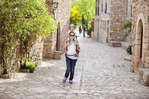 Young Caucasian woman tourist carrying her little son on shoulders while exploring old Spanish town