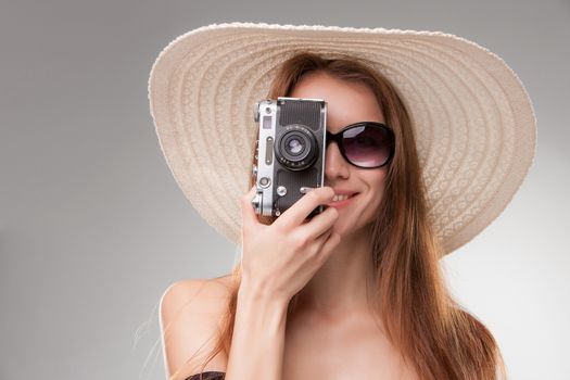 Girl in broad-brimmed hat and sunglasses with retro camera isolated on gray background