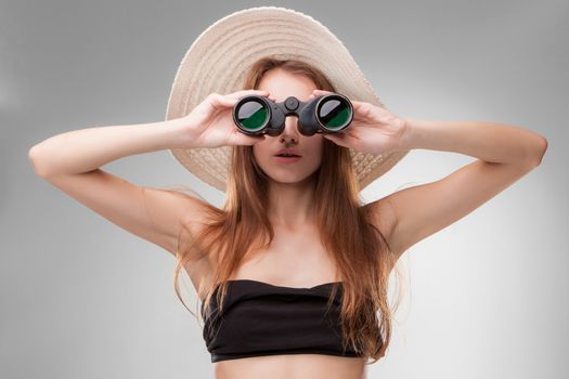 Young woman in hat with binoculars isolated on gray background. Travel and adventure concept. Closeup.