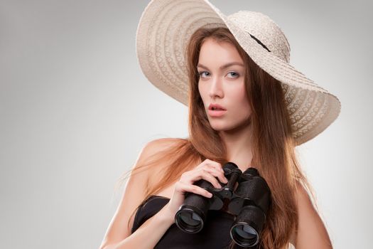 Young woman in hat with binoculars isolated on gray background. Travel and adventure concept. Closeup.