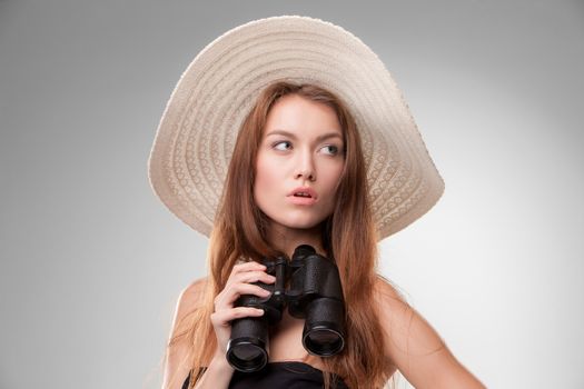 Young woman in hat with binoculars isolated on gray background. Travel and adventure concept. Closeup.