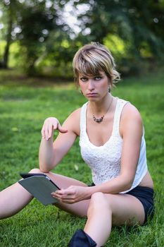 Portrait of a Gorgeous Young Woman with Short Blond Hair, Holding a Tablet Computer While Sitting on a Grassy Ground While Looking at the Camera.