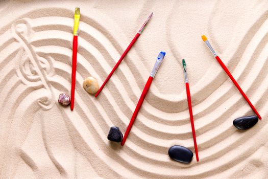 Harmony and music at the beach with an artistic conceptual image of a wavy score drawn in golden sand with a treble clef and musical notes of smooth pebbles and kids paintbrushes