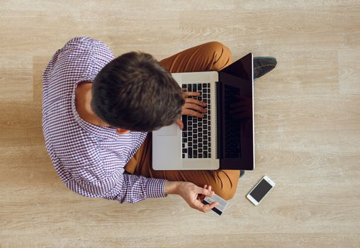 Online shopping - top view of young man sitting on the floor and working with a laptop and credit card in his hand
