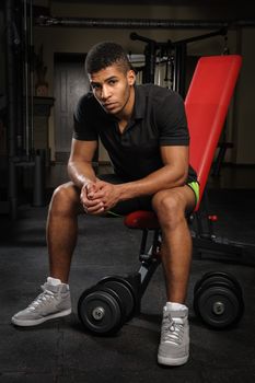 handsome young african descent mixed race man sitting and resting on bench in gym