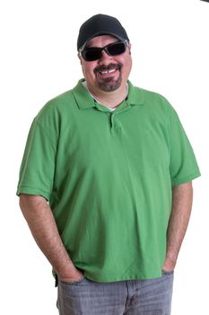 Portrait of Overweight Man Wearing Green Shirt, Baseball Cap, and Sunglasses Standing in Studio with Hands in Pockets of Jeans, in front of White Background and Smiling at Camera