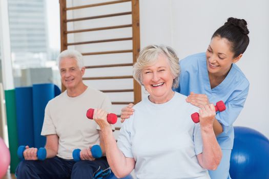 Portrait of happy senior woman lifting dumbbells while sitting with man and trainer in gym