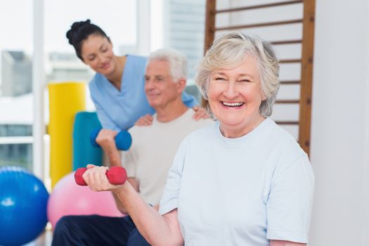 Portrait of happy senior woman lifting dumbbells while sitting with man and instructor in gym