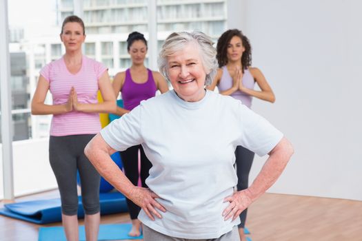 Portrait of happy senior woman with hands on hip standing against friends in gym