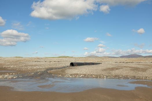 Water flows from a black drainage pipe down pebbles to a sandy beach area with hills and clouds in the distance.