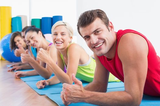 Portrait of happy man with friends gesturing thumbs up while lying on exercise mats at gym