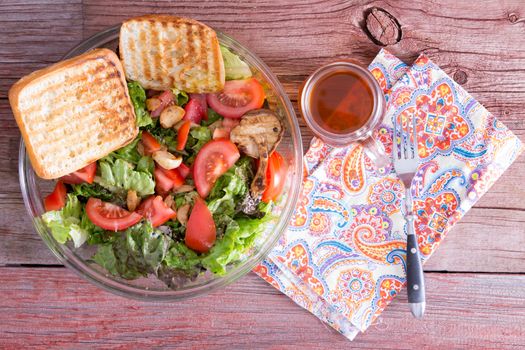 Healthy mixed leafy green salad with lettuce, tomato, garlic, sesame and bell peppers served with grilled toast and freshly brewed tea on a rustic wooden table and napkin, overhead view