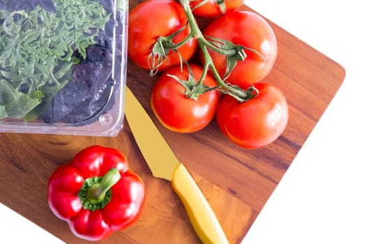 Healthy fresh salad ingredients on a chopping board with a sharp paring knife with three varieties of lettuve in a punnet and vine tomatoes with red bell pepper alongside, isolated on white