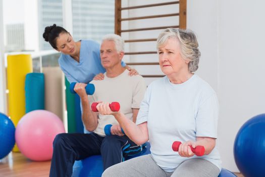 Senior woman lifting dumbbells with man and trainer in background at gym