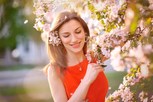 Young woman in red dress standing among blossom trees during sunny day  and wiping her nose. Girl with runny nose, having allergy and holding a tissue next to her face.