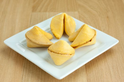 Four fortune cookies on a square white plate and sitting on a wooden table.