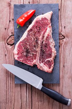 Close up Raw T-Bone Steak on Cutting Board with Salt, Whole Black Peppercorn and Knife on Top of a Wooden Table