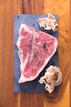 T-Bone Steak Meat Marinated with Whole Black Peppercorn and Golden Oak Shiitake Mushroom on Top of Gray Cutting Board on a Wooden Table.