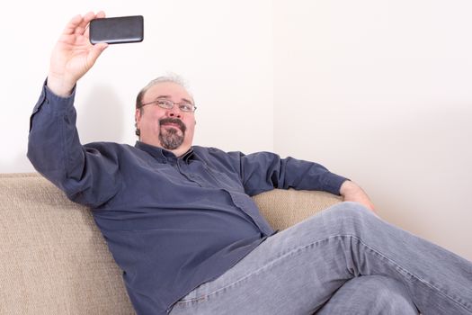 Middle-aged portly man wearing casual dark blue shirt and gray jeans while sitting on the sofa and taking selfie horizontal portrait pictures with a smart mobile phone, with copy space on white wall
