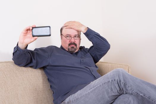 Vain man taking a selfie on his smartphone checking his hair with his hand as he looks critically at the screen while seated on a comfortable sofa at home