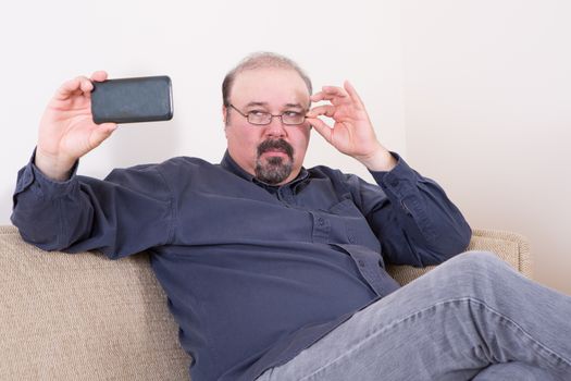 Fashionable middle-aged man taking a selfie on his mobile primping and adjusting his trendy glasses as he poses for the camera while sitting on a couch