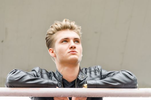 Attractive young man wearing black leather jacket looking outside from balcony