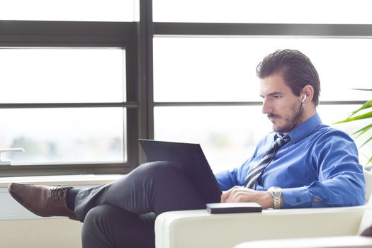 Busy, focused businessman in office working on his laptop wearing headphones. Side view. 