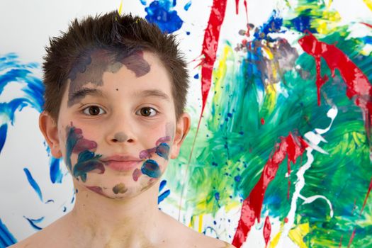 Handsome young boy daubed with colorful paint all over his face standing in front of his new modern abstract artwork with a blend of vibrant colors