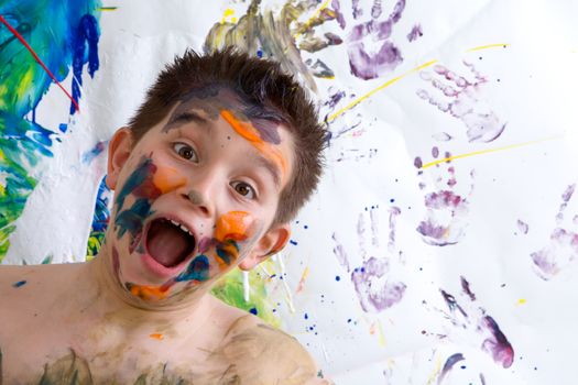 Excited happy little boy doing finger painting standing laughing at the camera with a wide open mouth in front of his artwork with vibrant colors and hand prints