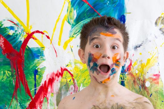 Astonished little boy with his face covered in colorful paint splodges gawping at the camera in front of a modern abstract painting in vibrant colors