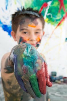 Artistic little boy holding out his paint covered hand to the camera as he stands in front of his modern abstract painting
