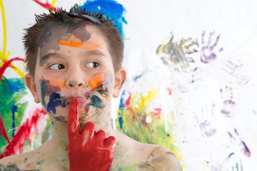 Thoughtful creative little boy covered in paint standing with his finger to his lips looking contemplatively to the side in front of his contemporary artwork as he seeks inspiration