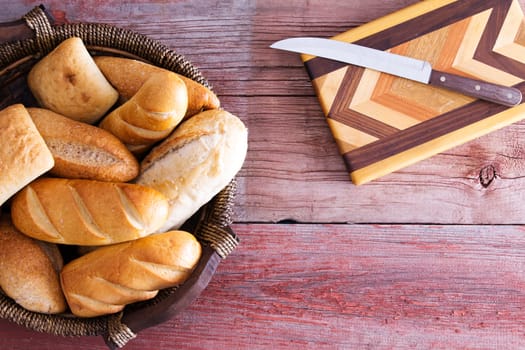 Assorted fresh rolls in a basket ready to be served for dinner standing on a rustic wooden table alongside a decorative inlaid wood cutting board and knife, overhead with copyspace