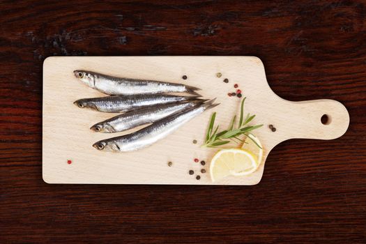 Delicious fresh sardines on wooden kitchen board with lemon, rosemary and colorful peppercorns on wooden background. Culinary healthy cooking.