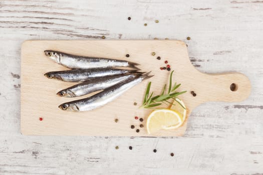 Delicious fresh sardines on wooden kitchen board with lemon, rosemary and colorful peppercorns on wooden background. Culinary healthy cooking.