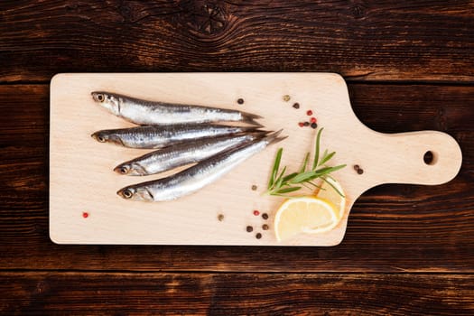 Delicious fresh sardines on wooden kitchen board with lemon, rosemary and colorful peppercorns on wooden background. Culinary healthy cooking.