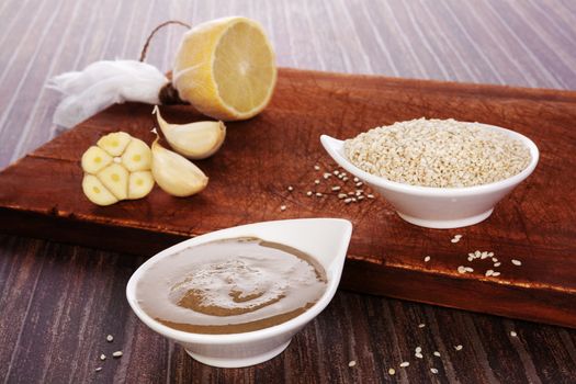 Tahini pasta in bowl, sesame seeds and lemon on brown wooden background. Culinary eating. 