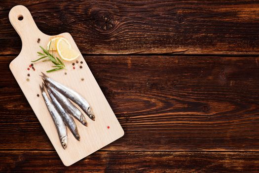 Delicious fresh sardines on wooden kitchen board with lemon, rosemary and colorful peppercorns on wooden background with copy space. Culinary healthy cooking.