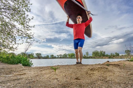 senior male paddler carrying his SUP paddleboard on a lake shore in Colorado, early spring