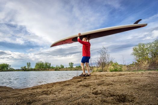 senior male paddler carrying his SUP paddleboard on a lake shore in Colorado, early spring