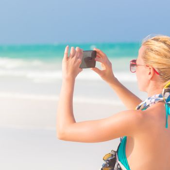 Blonde caucasian woman taking photo of blue tropical beach. Beautiful caucasian model  wearing turquoise swimsuit and colorful scarf on vacations on picture perfect Paje beach, Zanzibar, Tanzania.