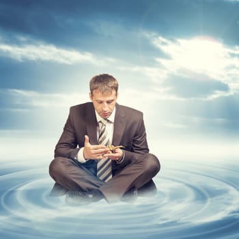 Businessman in suit sitting in lotus position on cloud with circles and looking at oil lamp on blue sky background
