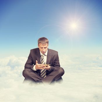 Businessman in suit sitting in lotus position on cloud and looking at oil lamp on blue sky background