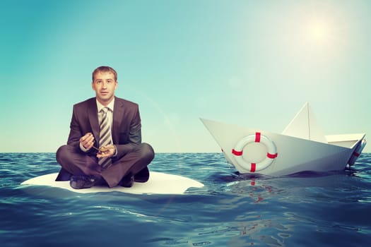 Businessman sitting in lotus position with paper boat in sea and looking at camera