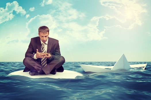 Businessman sitting in lotus position on small sand island in sea and looking up, sinking paper boat on right side