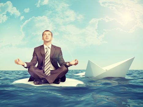 Businessman sitting in lotus position on small island in sea and looking up, sinking paper boat on right side
