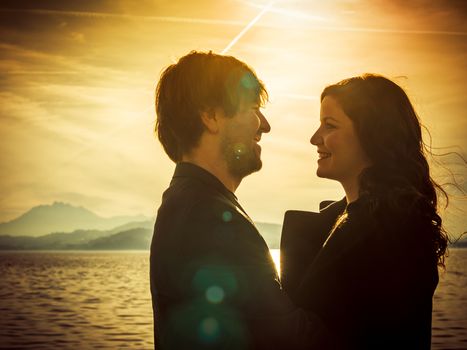 Photo of young adults by the water enjoying the warmth of the sun in the late afternoon in autumn. Heavily filtered for more romantic effect. Real lens flare visible.