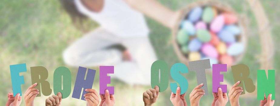 Hands holding up frohe ostern against little girl sitting on grass showing basket of easter eggs to camera