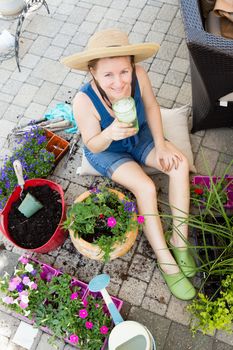 Attractive woman potting up houseplants in spring sitting on a brick patio surrounded by plants, fertile soil and containers enjoying a refreshing glass of iced water in the sunshine, view from above
