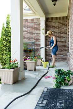 Woman gardener standing watering newly potted decorative spring plants arranged in flowerpots along an open-air patio using a hosepipe and sprinkler spray